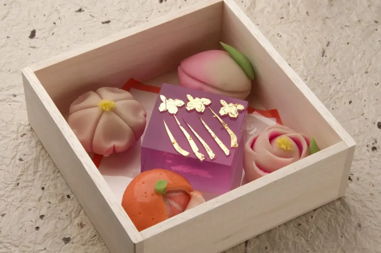 Schachtel mit kunstvollen japanischen Desserts: Blüten, Pfirsich, Rose und eine orangefarbene Frucht, verziert mit Blattgold.