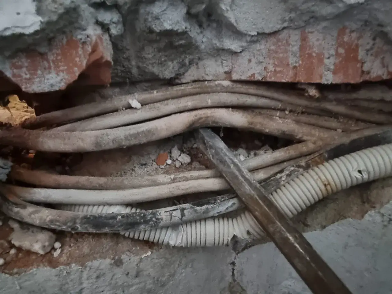Uszkodzone przewody elektryczne w ścianie, grożące zwarciem i pożarem. Widać fragmenty cegły i tynku.