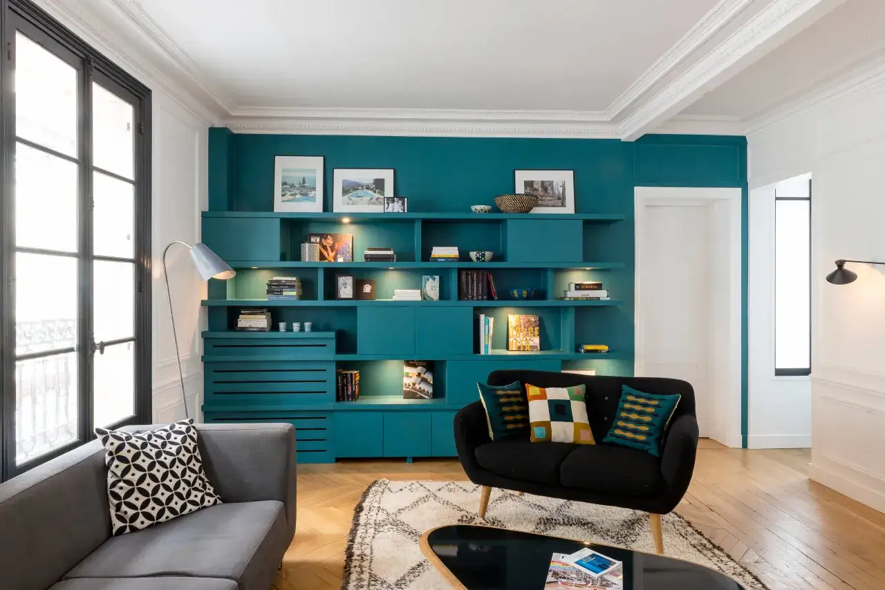 Comment éclaircir une pièce sombre : un salon moderne avec un mur bibliothèque bleu canard, un canapé gris, un fauteuil noir et un grand lustre.