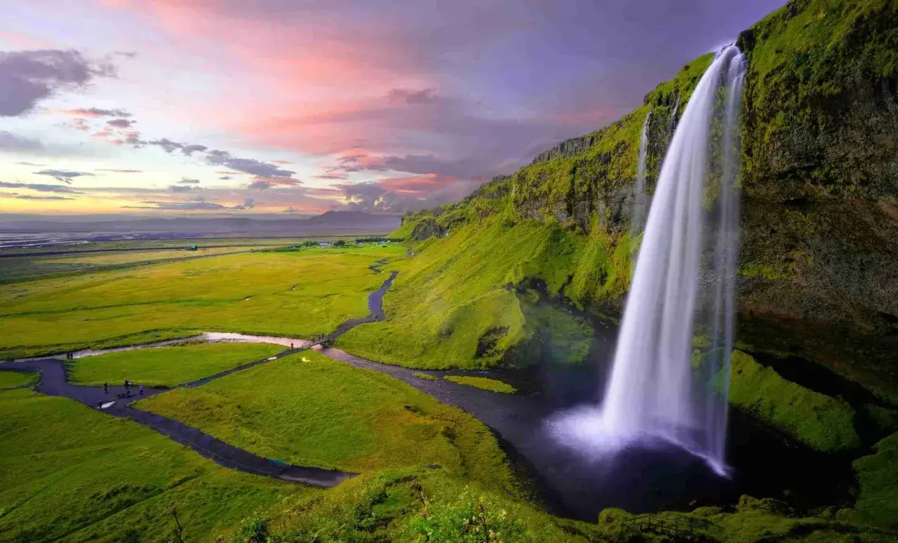 Ein majestätischer Wasserfall stürzt von einer moosbewachsenen Klippe in ein dunkles Becken. Ein Fluss schlängelt sich durch eine grüne Ebene, ideal für einen Roadtrip auf dieser Insel.
