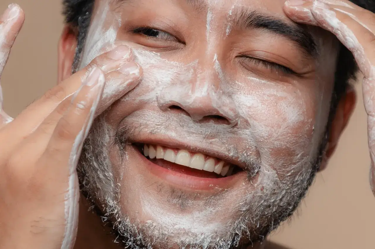 Un homme souriant applique un masque blanc sur son visage, une &eacute;tape cl&eacute; pour savoir comment enlever les boutons et obtenir une peau nette.