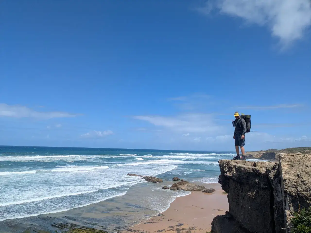 Wanderer mit Rucksack steht auf Klippe in Aljezur, Portugal, blickt auf tosendes Meer und einsamen Strand.