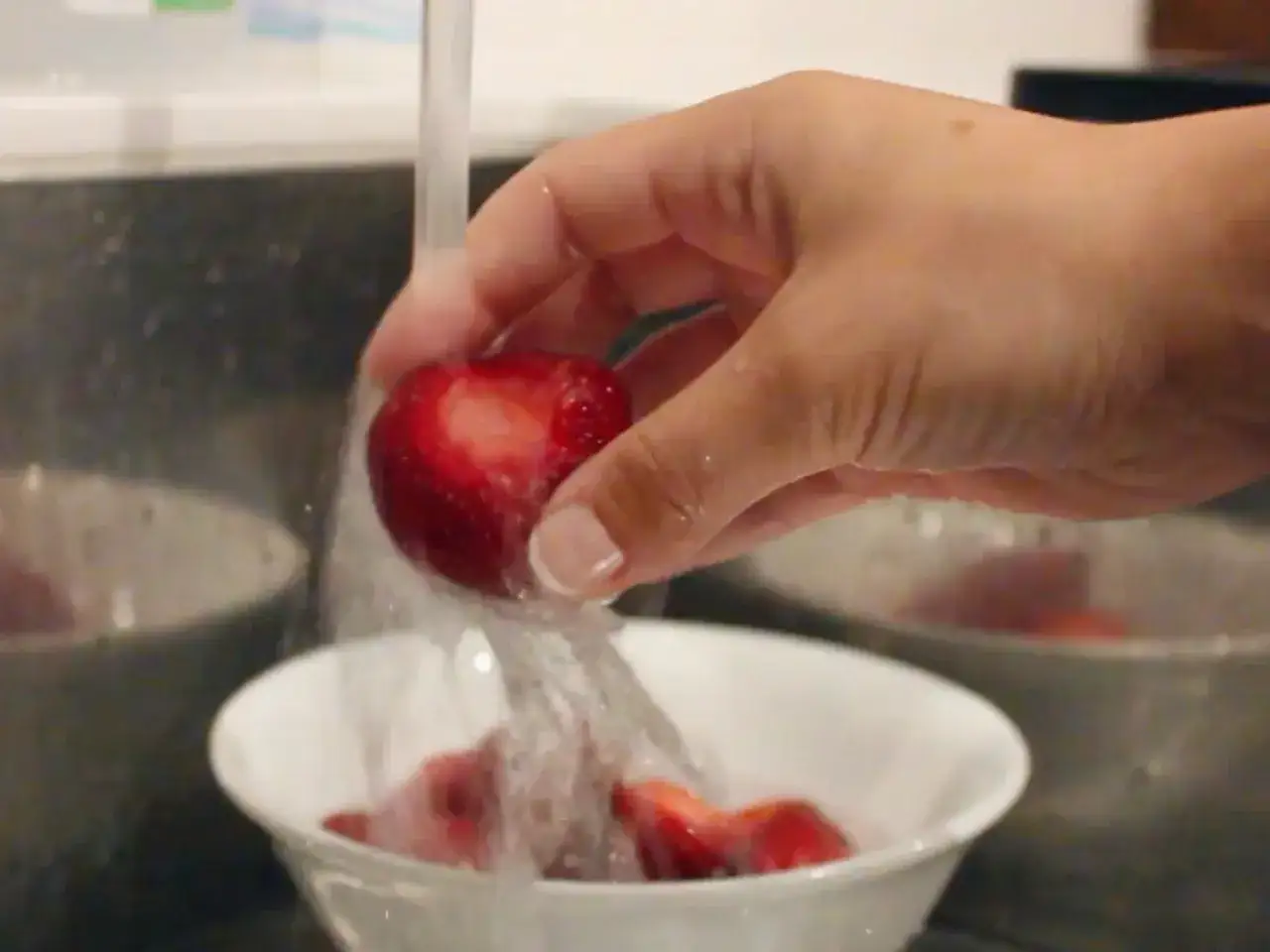 Eine Hand hält eine rote Erdbeere unter fließendem Wasser, um sie in einer weißen Schüssel zu waschen.