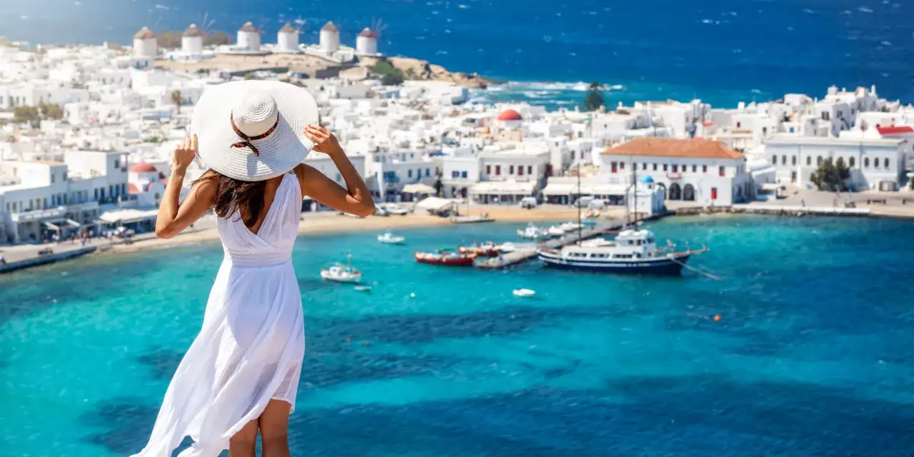 Kobieta w białej sukience i kapeluszu podziwia widok na białe domki i wiatraki na Mykonos. Czy w Grecji jest drogo? Widok zapiera dech!
