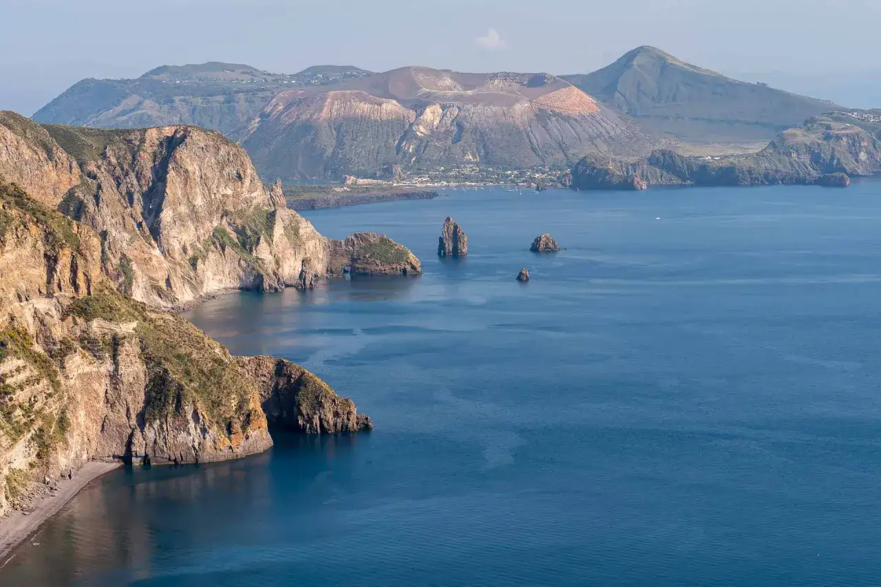 Vue panoramique de l'&icirc;le de Lipari, avec ses falaises rocheuses escarp&eacute;es plongeant dans la mer bleue et un volcan imposant en arri&egrave;re-plan.