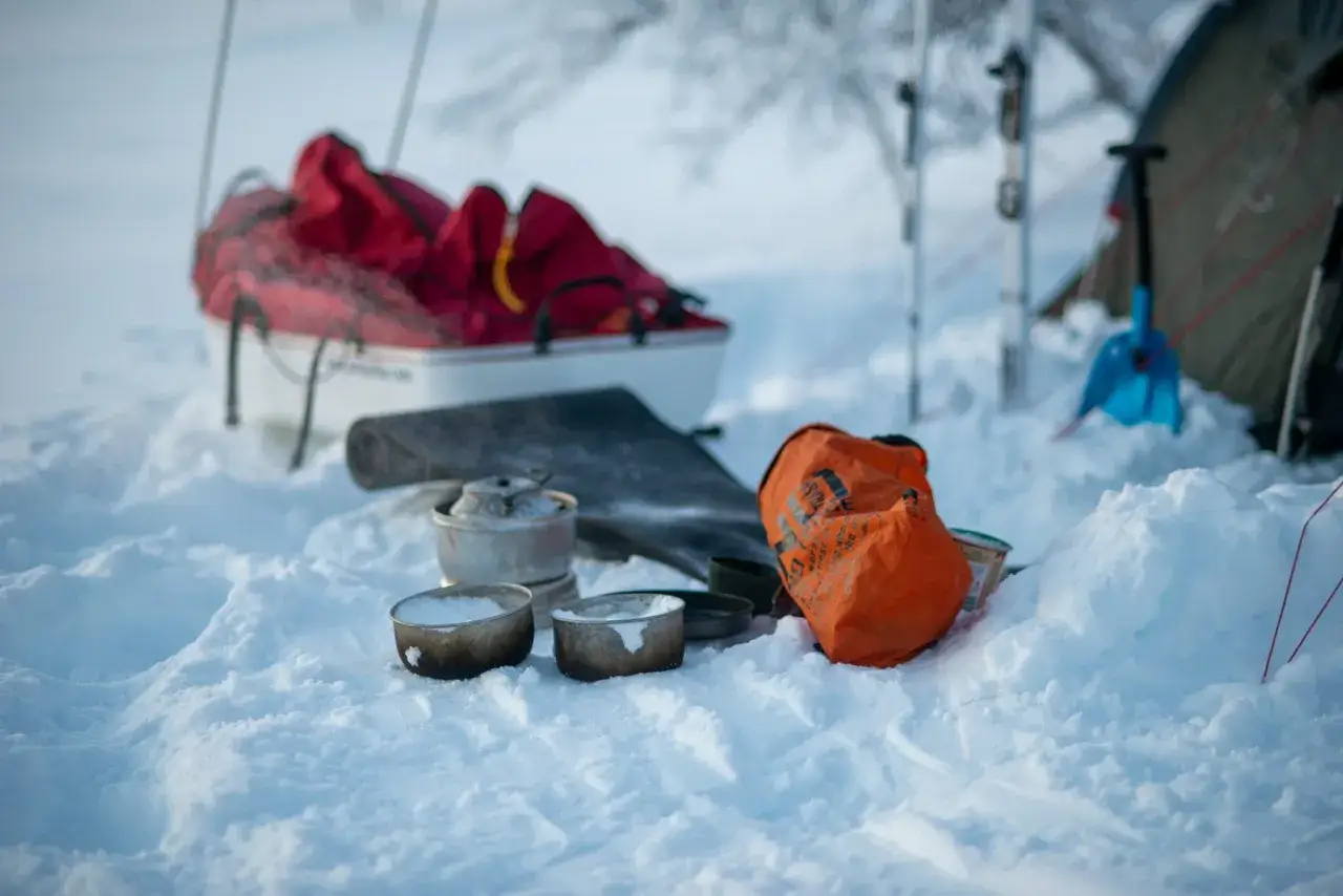 Vinter camping-utrustning i snön: kastrull, skålar, orange väska, släde med röda packpåsar, tält och skidor.