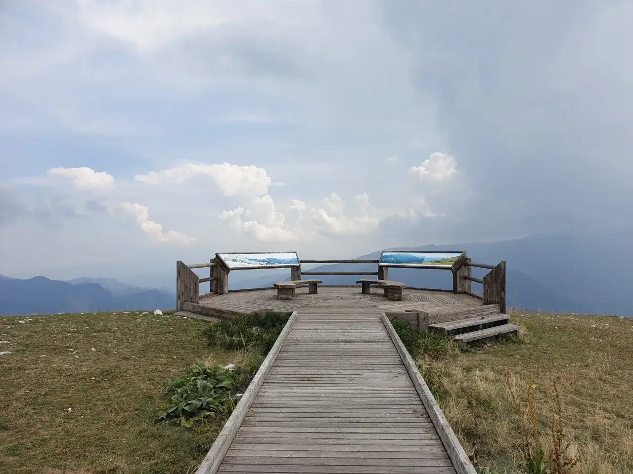 Terrazza panoramica in legno a Forca di Presta, con panchine e pannelli informativi, affacciata su un paesaggio montano nebbioso.