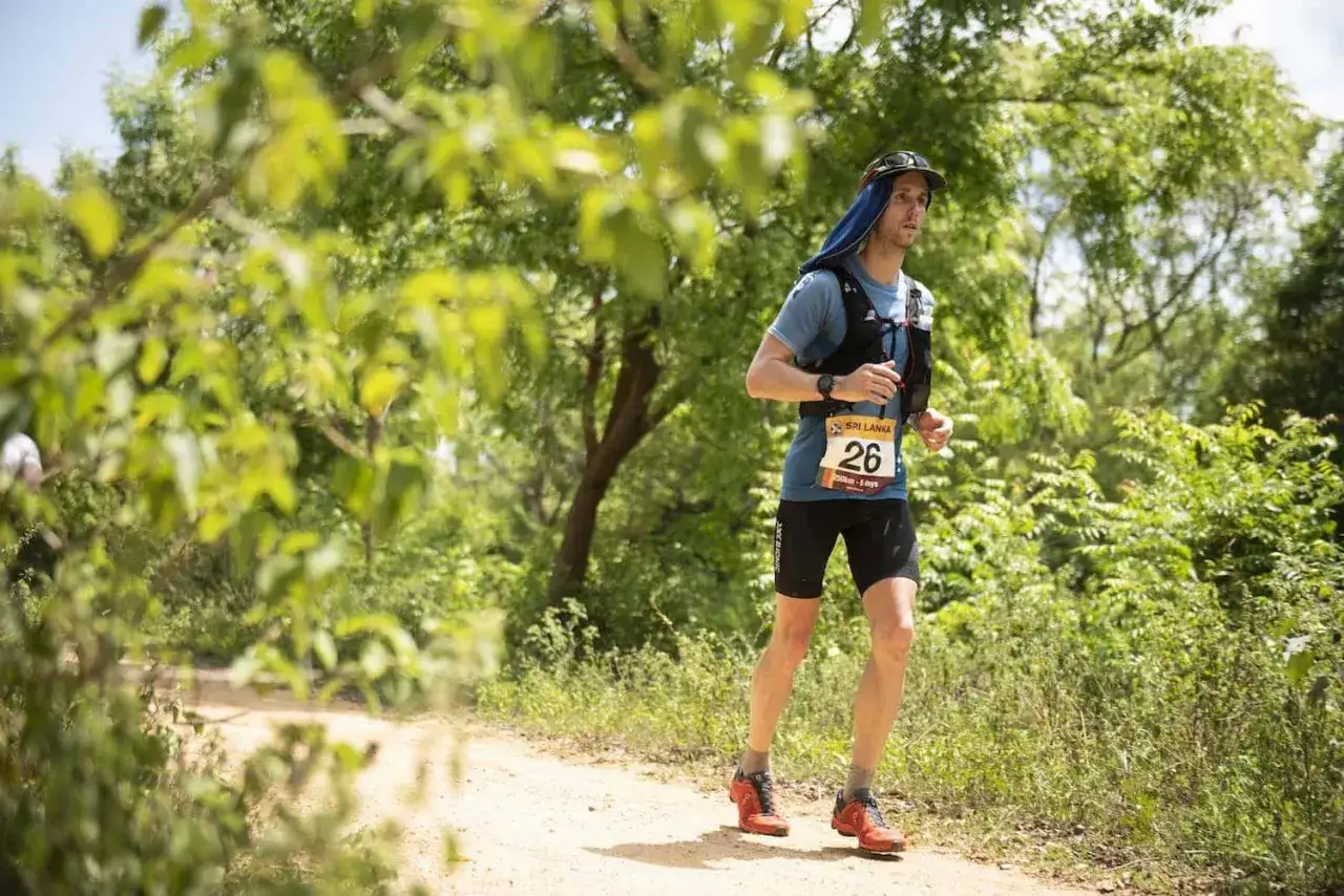 Atleta con pettorale 26 corre su un sentiero sterrato, parte di una corsa a tappe in un ambiente verdeggiante.