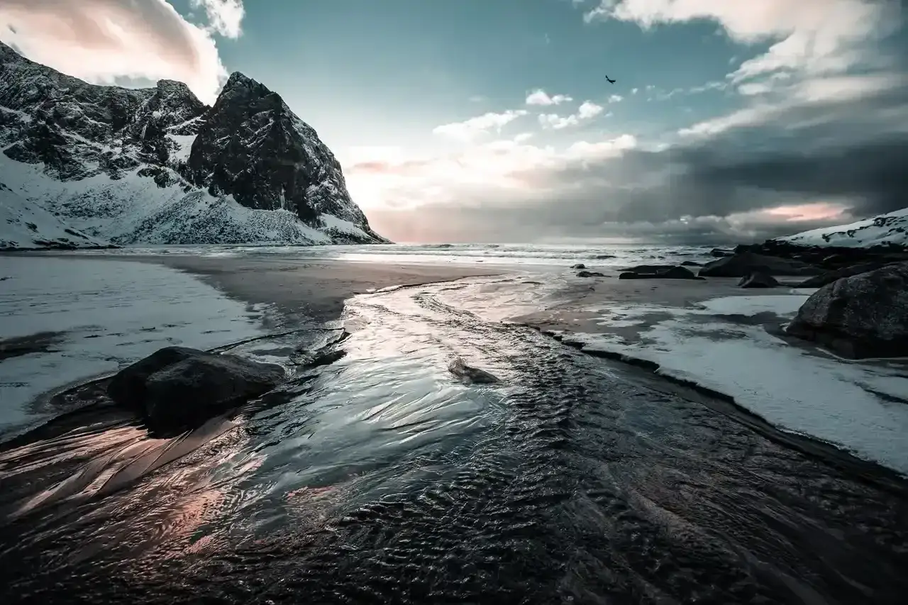 Zimowy strumyk płynie ku morzu na tle ośnieżonych gór. Idealne miejsce na norwegia wakacje.