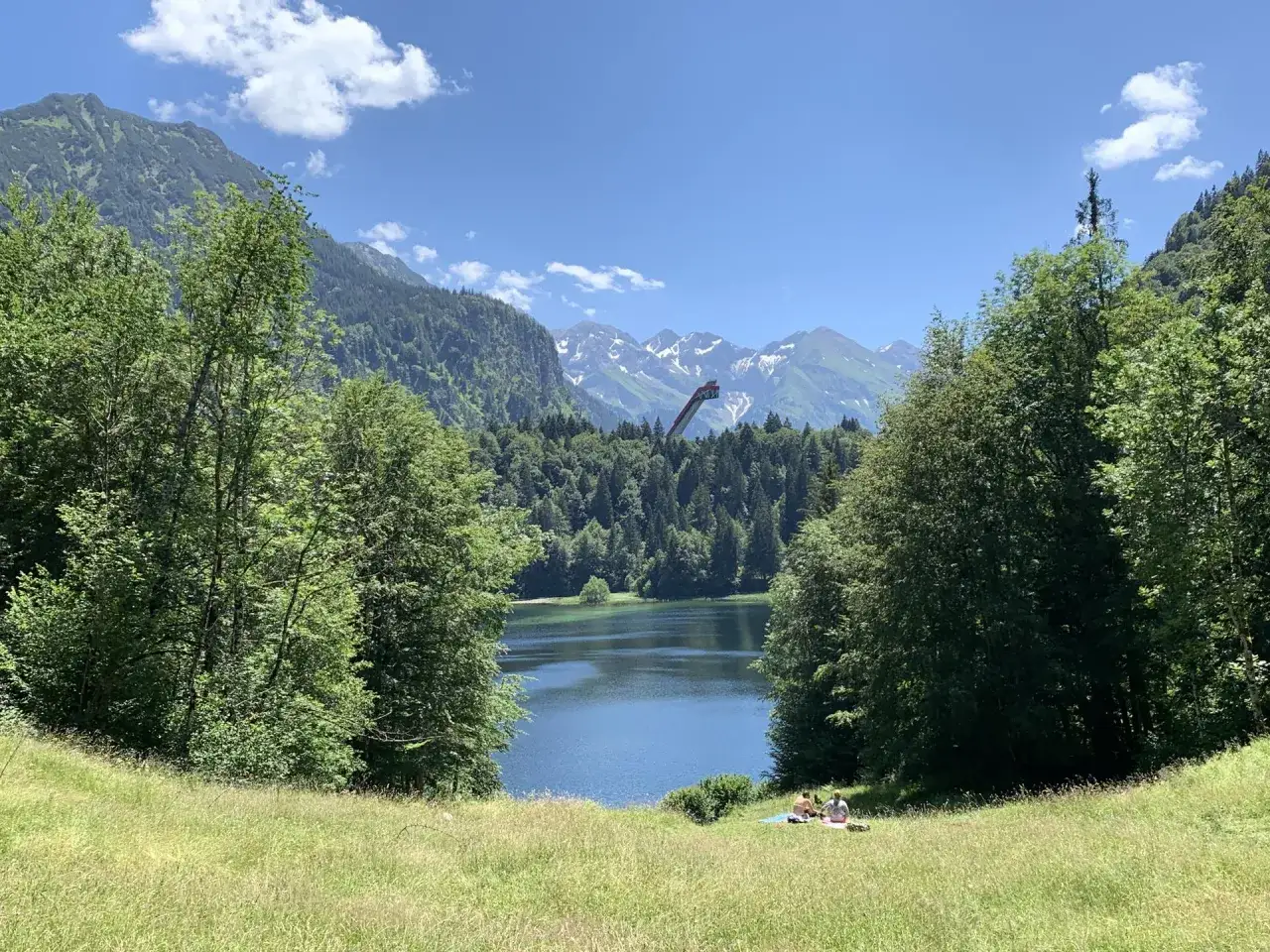 Idyllischer Sonthofener See mit schneebedeckten Berggipfeln im Hintergrund. Zwei Personen genie&szlig;en ein Picknick auf einer sonnigen Wiese.