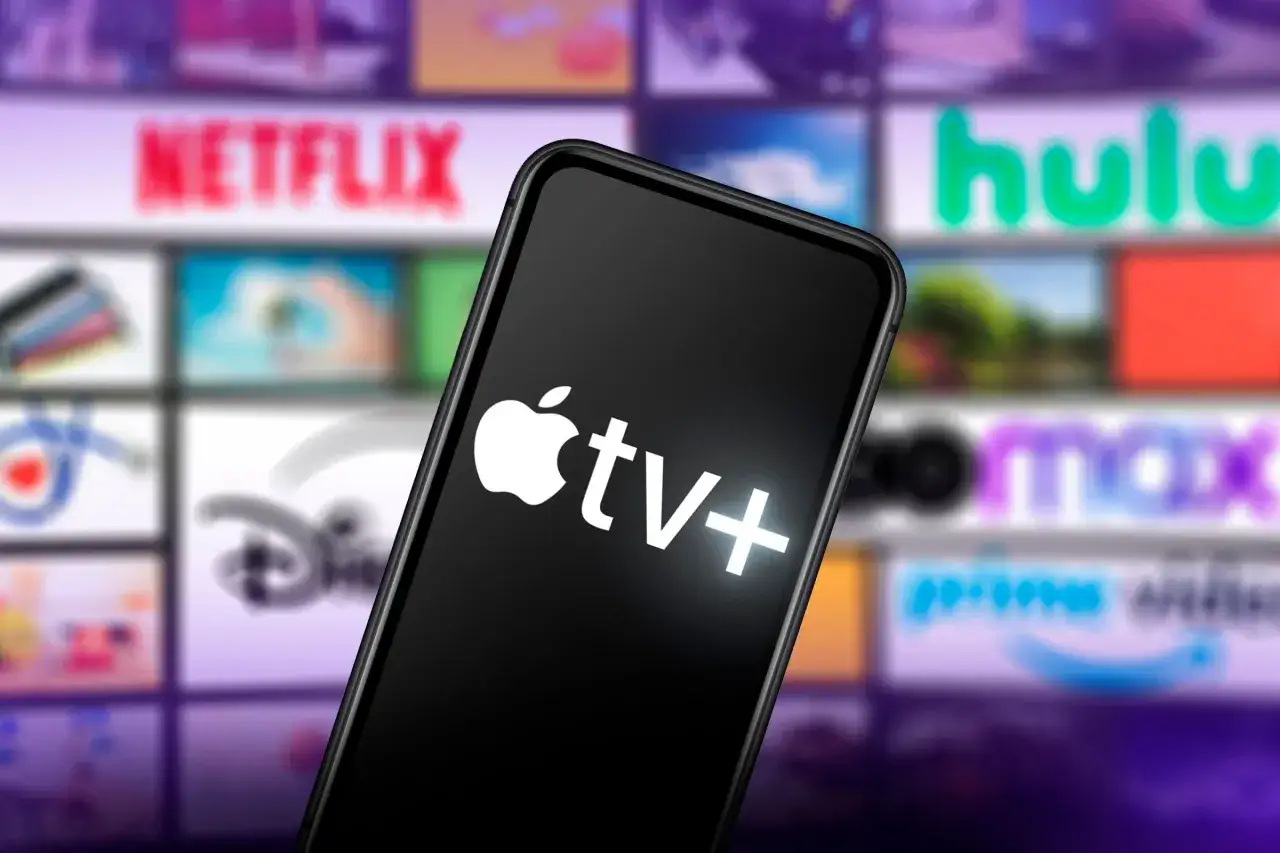 Smartphone zeigt das Apple TV+ Logo vor einer Collage aus Streaming-Diensten wie Netflix und Hulu.