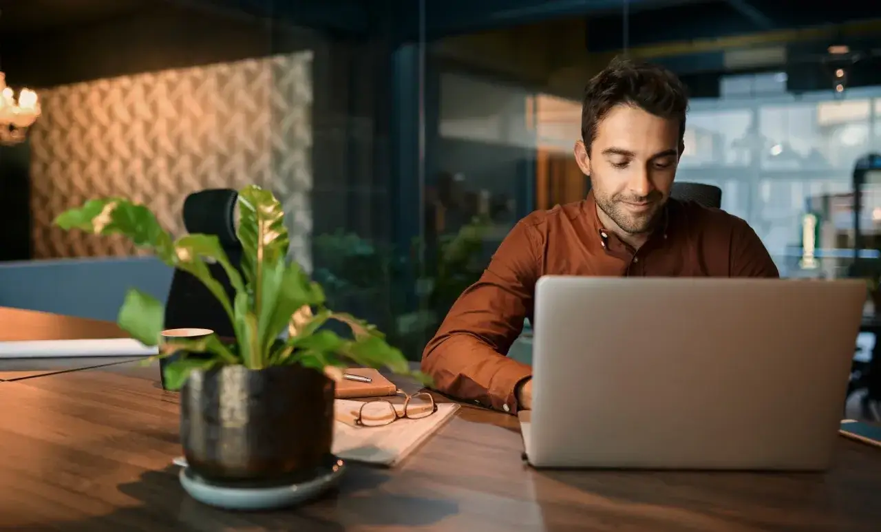 Man med brun skjorta jobbar vid laptop. En krukv&auml;xt och glas&ouml;gon ligger p&aring; skrivbordet. K&auml;nns som en mygla p&aring; jobbet.