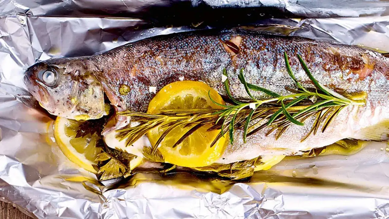 Ein ganzer Fisch, zubereitet als Forellenfilet im Backofen, liegt auf Alufolie mit Zitronenscheiben und Rosmarin.