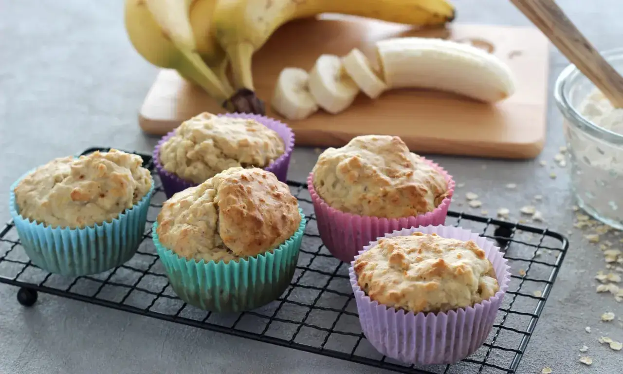 F&uuml;nf leckere bananenmuffins ohne zucker in bunten F&ouml;rmchen auf einem Gitter. Bananen und geschnittene Bananen liegen im Hintergrund.
