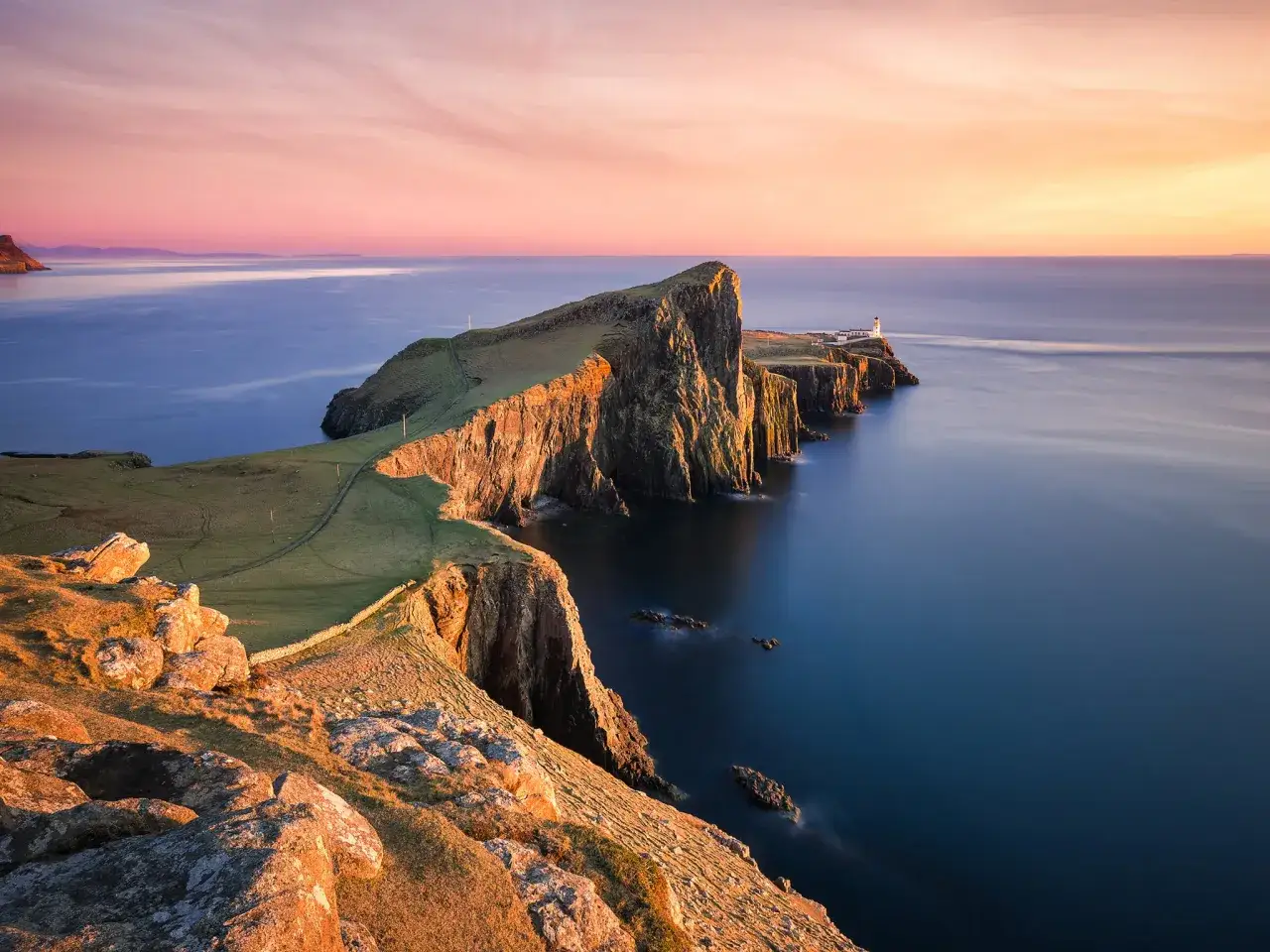 A lighthouse stands on a dramatic cliff edge, overlooking the sea. Winding hiking trails in Scotland lead across the grassy headland, bathed in the warm glow of sunset.