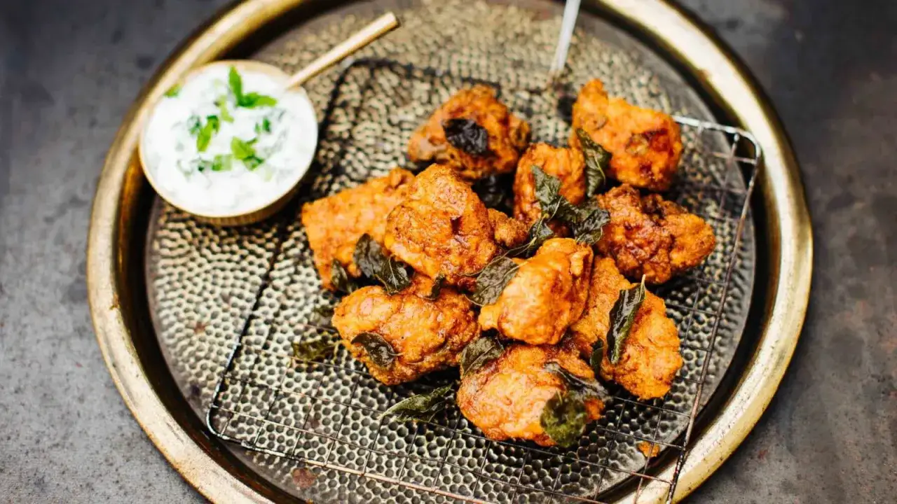 Knusprige chicken pakora auf einem Gitter, garniert mit frischen Kräutern, daneben eine Schale Joghurt-Dip.