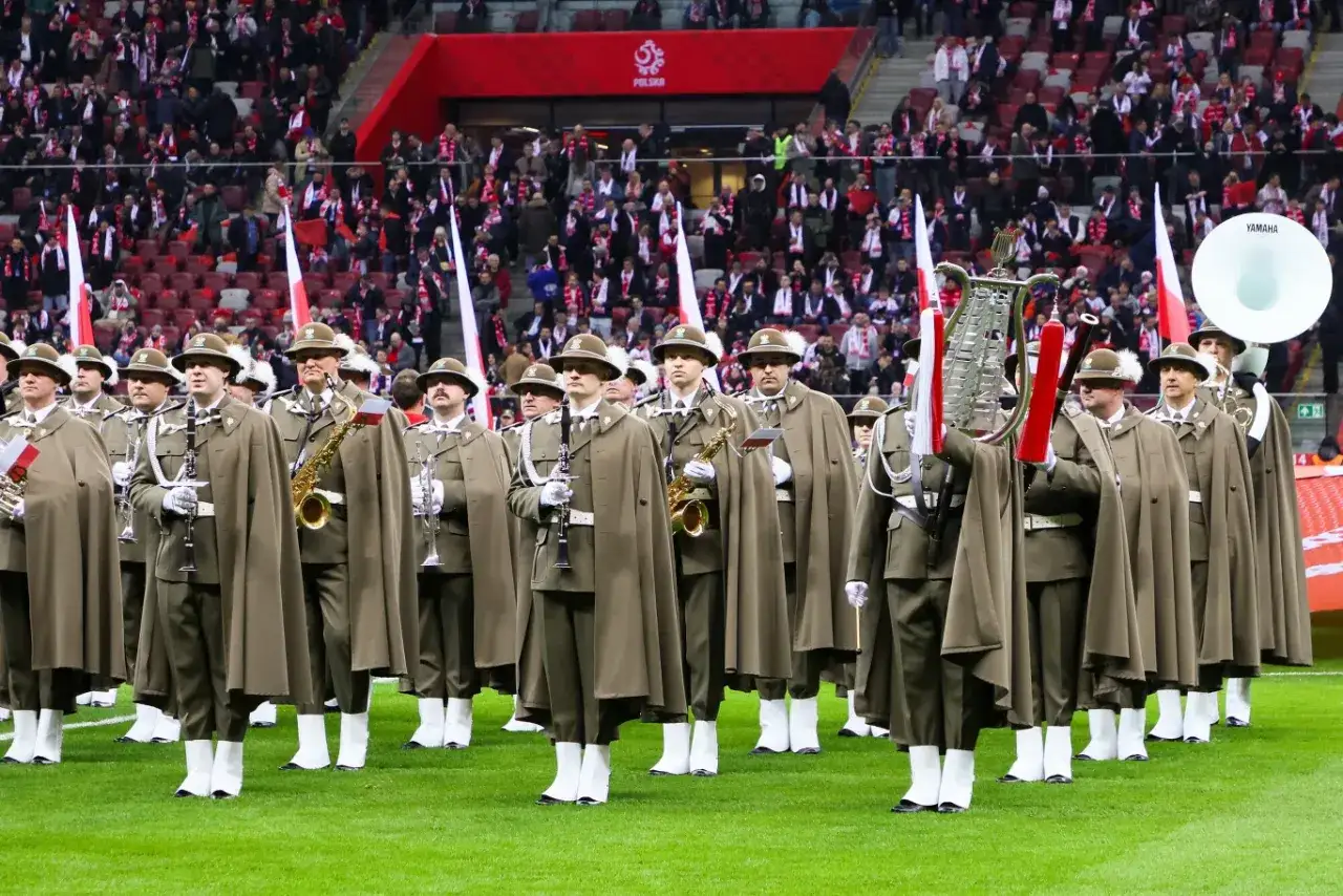 Orkiestra wojskowa w strojach galowych, gotowa do zagrania hymnu. W tle trybuny stadionu wypełnione kibicami.