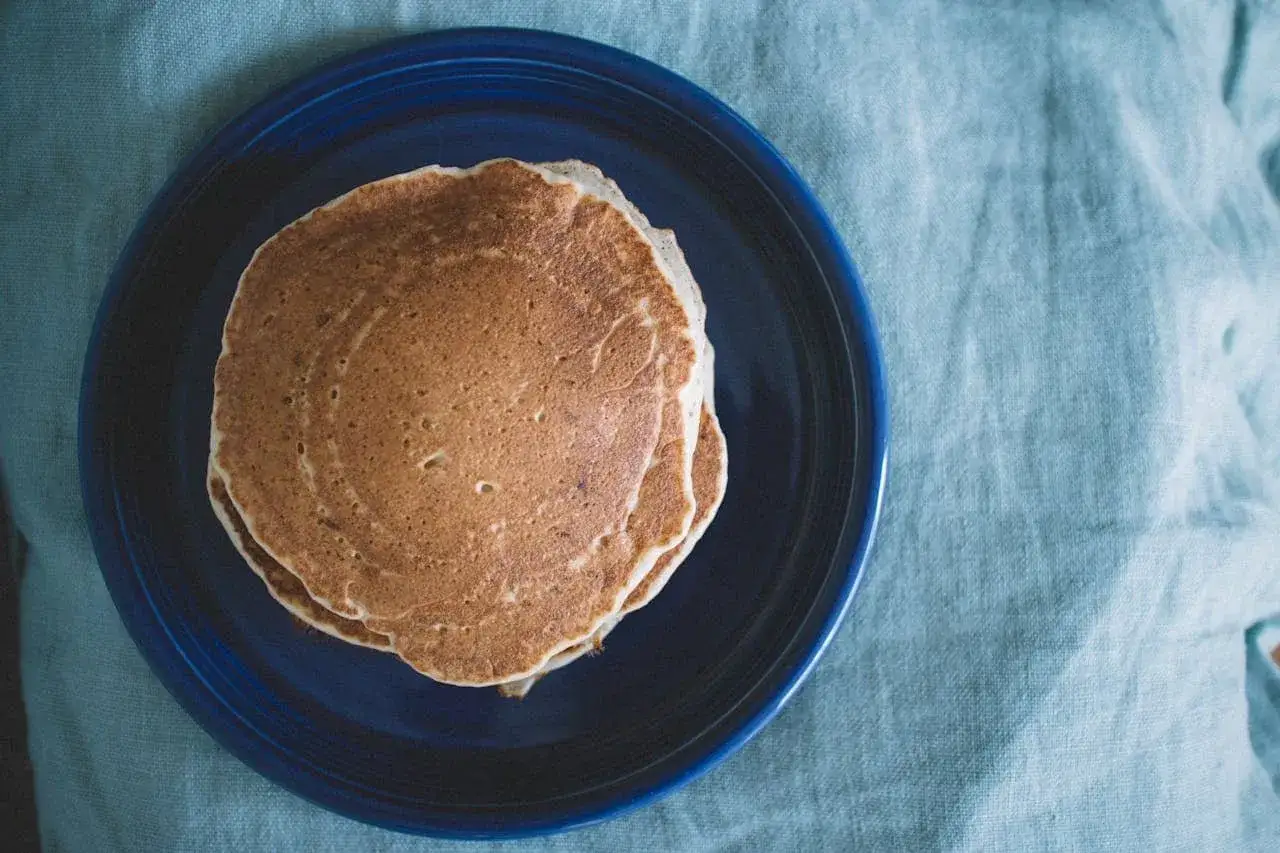 Pile de pancakes au levain dorés sur une assiette bleue, posée sur un tissu bleu clair froissé.