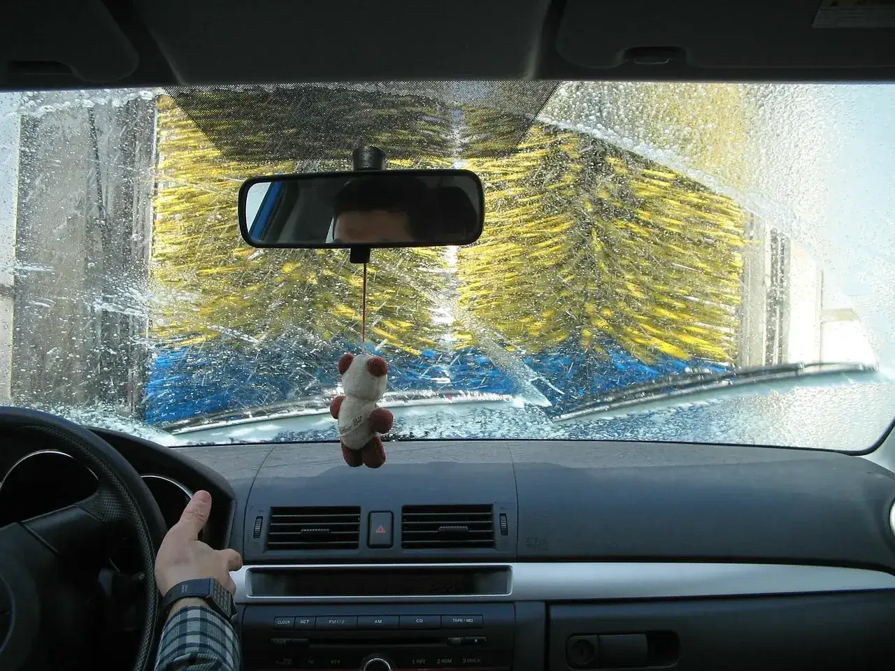 Widok z wnętrza samochodu podczas mycia. Widać wielkie, żółte szczotki myjni automatycznej i krople wody na szybie.