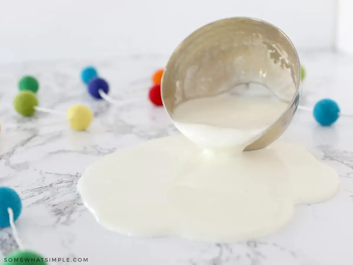 Ein umgekippter Becher mit weißer Flüssigkeit, die sich auf Marmor ausbreitet. Bunte Pompons im Hintergrund deuten auf ein lustiges Oobleck-Rezept hin.