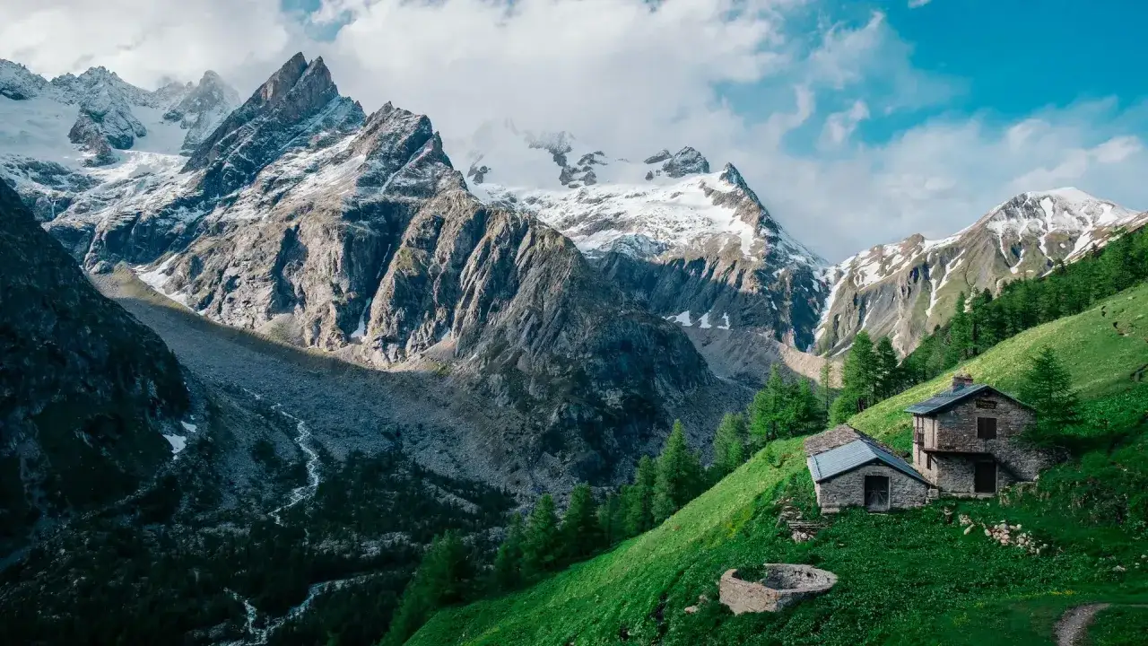 Stone huts dot a lush green slope, a picturesque stop on a self-guided tour du Mont Blanc, with dramatic snow-capped peaks looming behind.