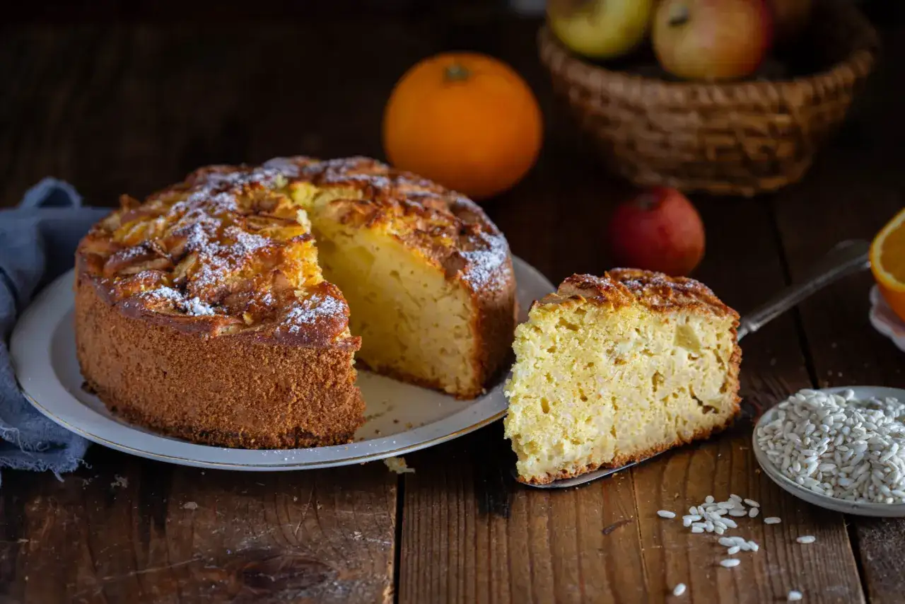 Torta soffice con farina di riso, guarnita con mele e zucchero a velo, accanto a un piatto di riso crudo.