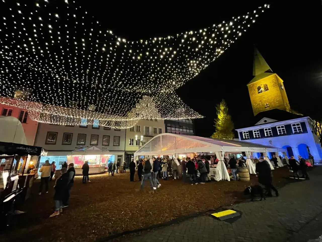 Glitzernde Lichterketten überspannen den stimmungsvollen weihnachtsmarkt in Dorsten. Menschen bummeln zwischen Ständen und Zelten, während im Hintergrund die beleuchtete Kirche thront.