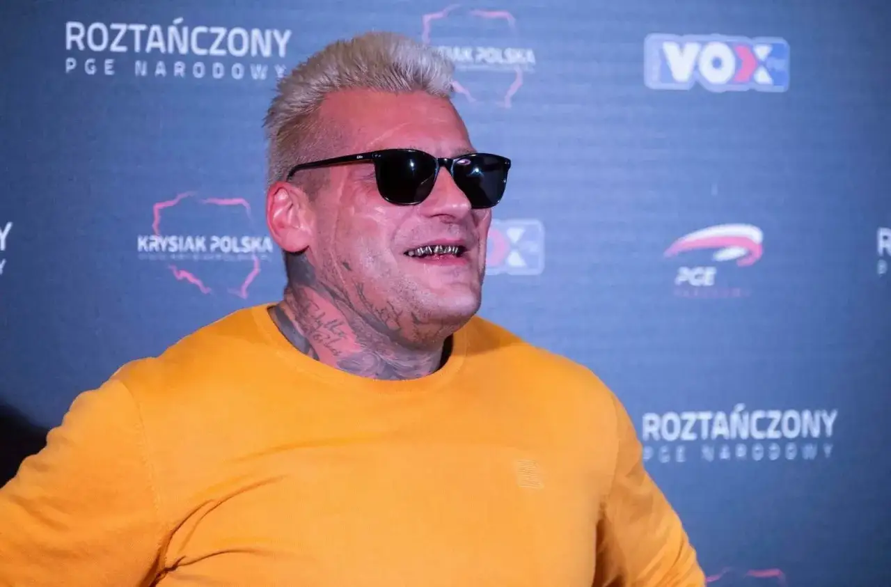 Popek kiedys, z charakterystycznymi złotymi zębami i tatuażami, w pomarańczowej bluzie i okularach przeciwsłonecznych, na tle napisu