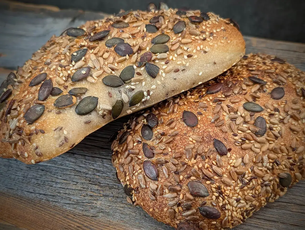 Zwei rustikale Ciabatta Brote, bedeckt mit Sonnenblumenkernen, K&uuml;rbiskernen und Sesam, liegen auf einer Holzoberfl&auml;che.