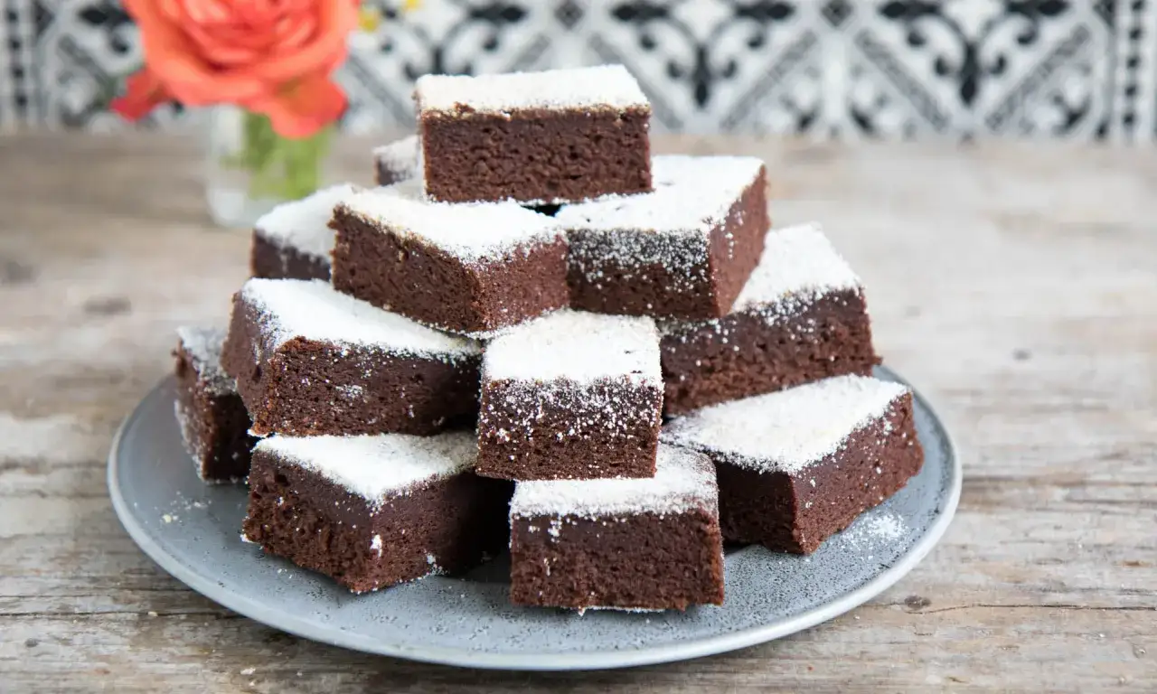Ein Stapel saftiger Brownies mit Datteln, best&auml;ubt mit Puderzucker, auf einem grauen Teller.