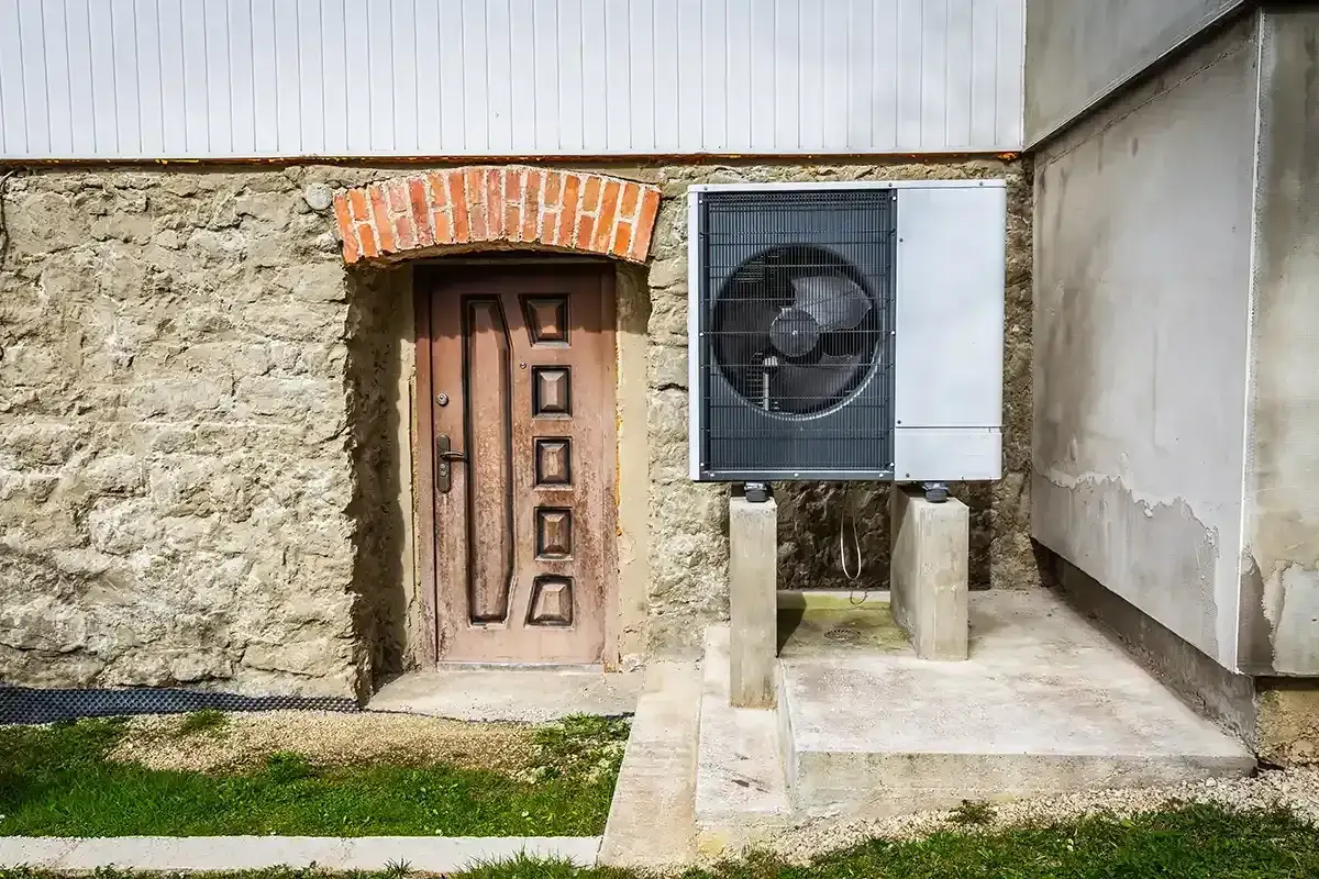 Nowoczesna pompa ciepła do starej instalacji grzewczej, zamontowana obok kamiennego muru i drewnianych drzwi.
