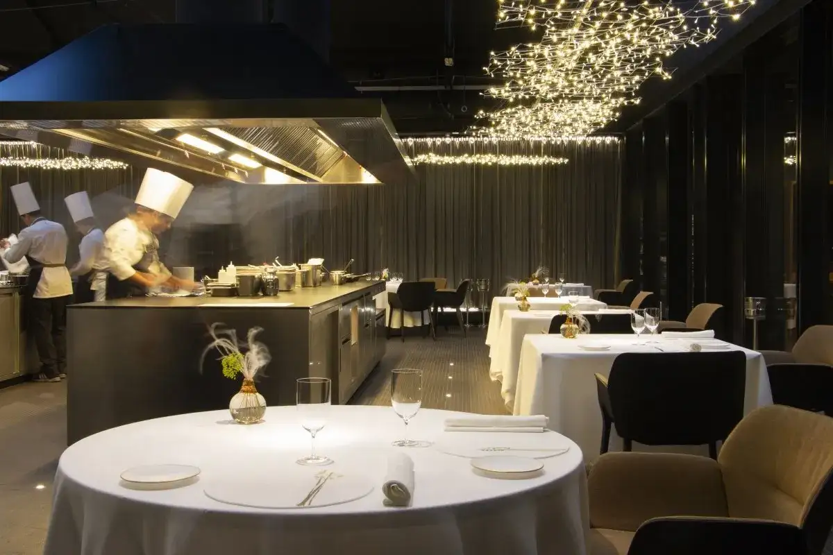 Chefs trabajan en una cocina abierta de un restaurante con **4 estrellas Michelin**. Mesas elegantemente puestas esperan a los comensales bajo una cascada de luces.