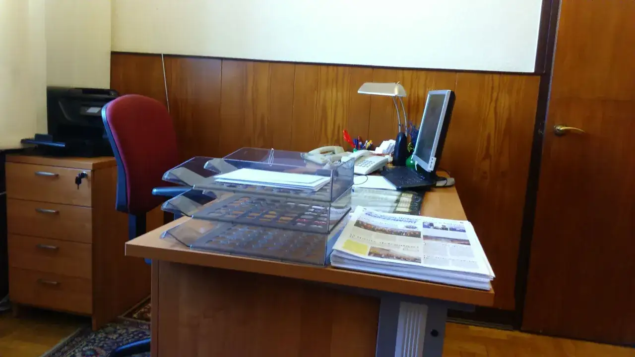 Biurko z komputerem, telefonem, lampką i stosem gazet. Obok drukarka i czerwone krzesło. To miejsce pracy, gdzie realizowane są obowiązki pracownika.