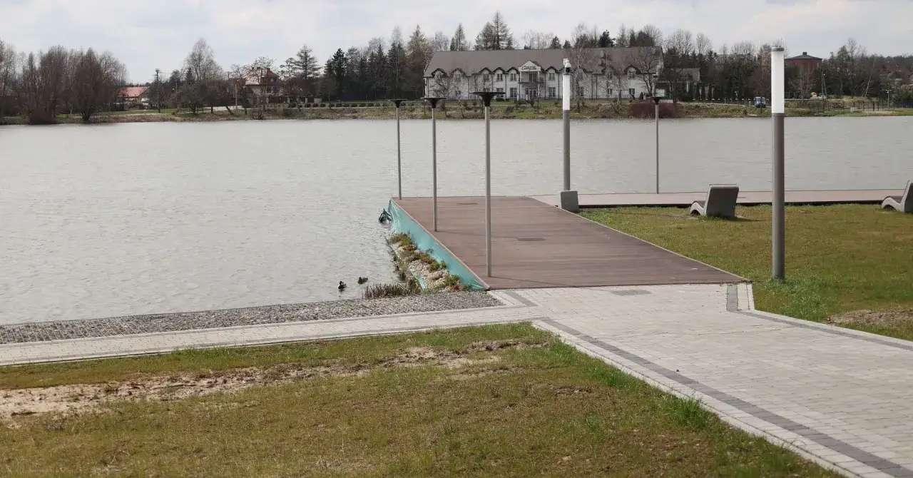 Drewniane molo nad zalewem wolbromskim, z kaczkami pływającymi w wodzie i budynkiem w tle.