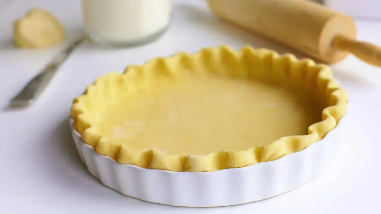P&acirc;te bris&eacute;e pr&ecirc;te &agrave; cuire dans un moule blanc. Le temps de cuisson p&acirc;te bris&eacute;e est crucial pour une tarte r&eacute;ussie. Rouleau &agrave; p&acirc;tisserie en arri&egrave;re-plan.