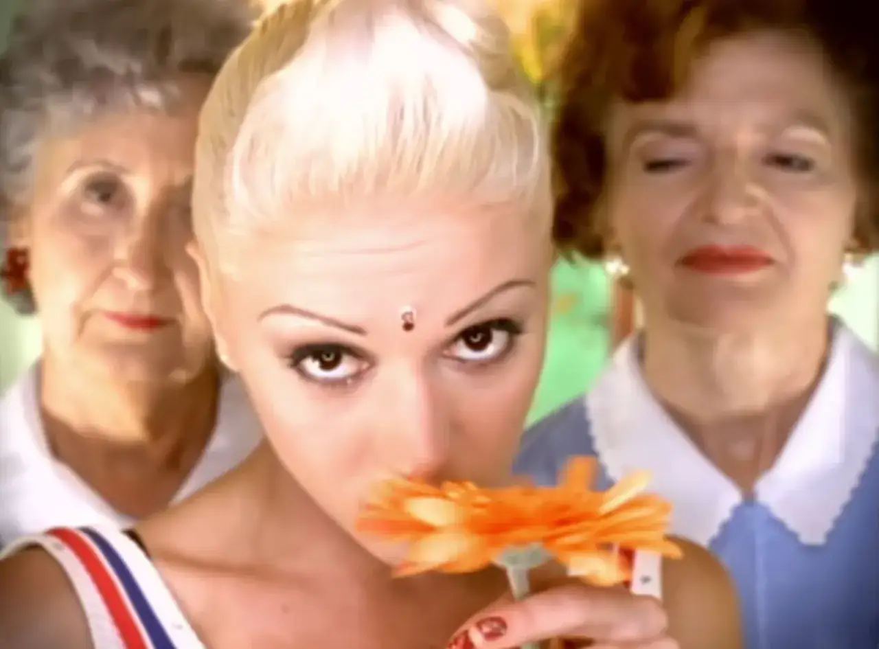 Gwen Stefani wącha pomarańczową gerberę, podczas gdy dwie starsze kobiety patrzą zza niej.