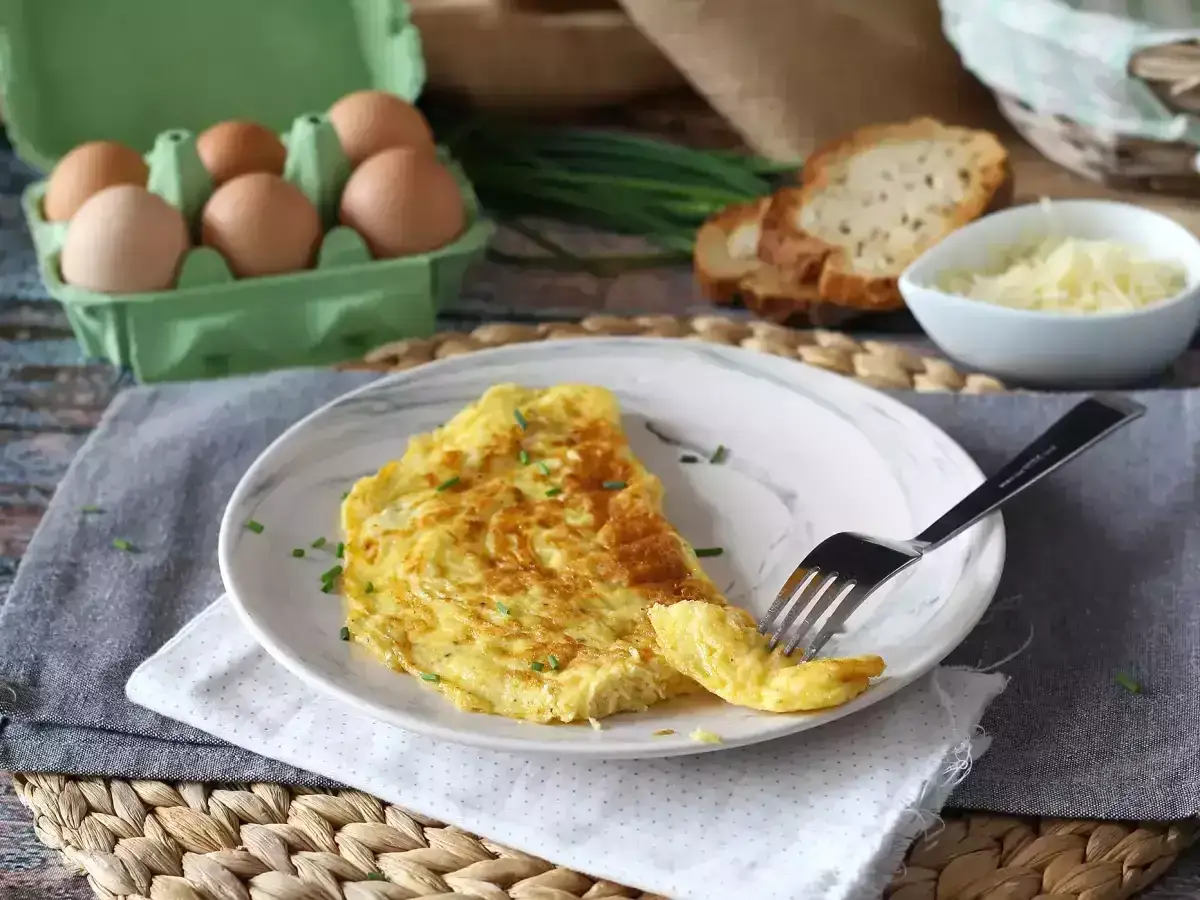 Puszysty omlet francuski, posypany szczypiorkiem, czeka na widelcu. Obok kromki chleba, starty ser i jajka.