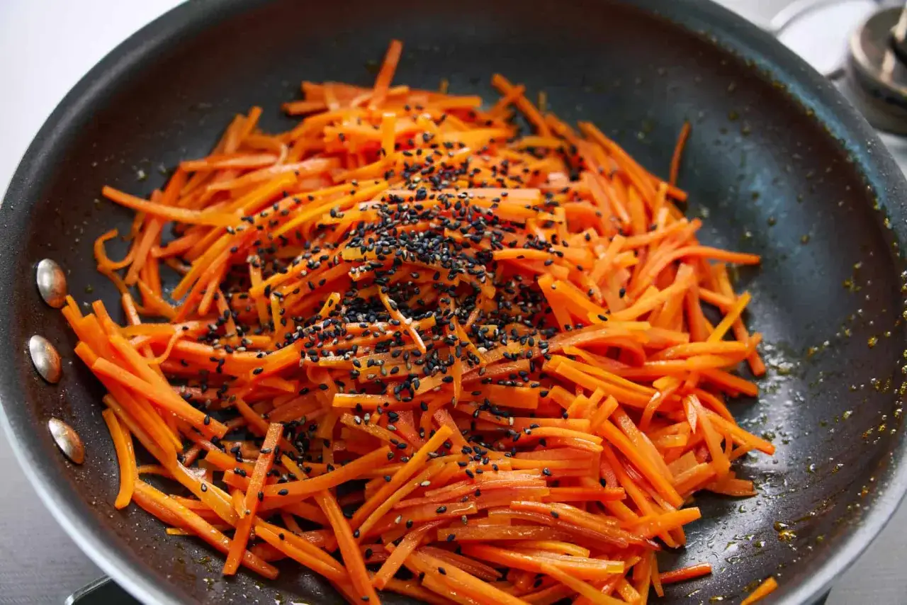 Shredded carrots are being stir-fried in a pan, sprinkled with black sesame seeds, creating a vibrant dish of Japanese pickled carrots.