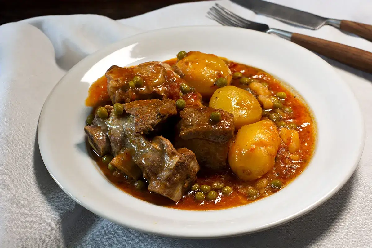 Plato de patatas con costillas estofadas en salsa roja, con guisantes.