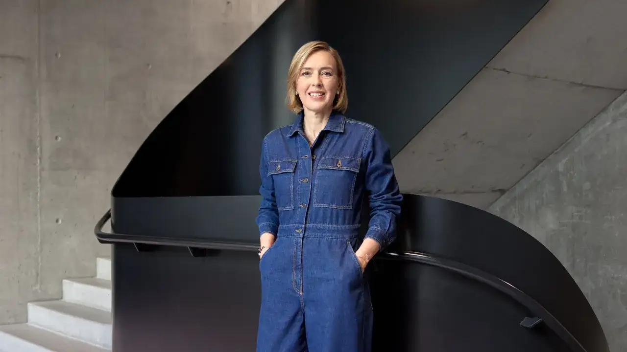 Christiane von Hardenberg l&auml;chelt in einem Jeans-Overall, die H&auml;nde in den Taschen, vor einer modernen Treppe aus Beton und schwarzem Metall.