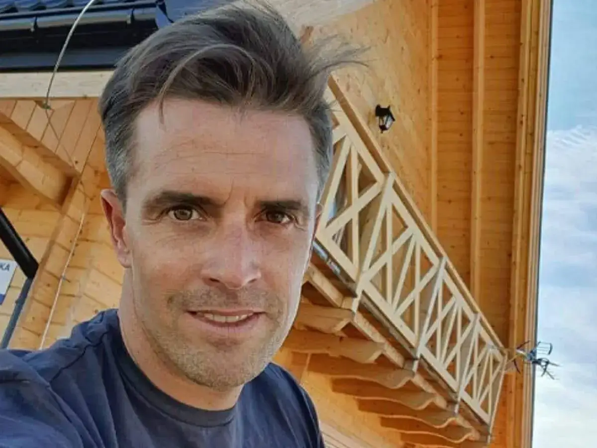 Staszek Karpiel-Bułecka uśmiecha się przed drewnianym domem z charakterystyczną balustradą.