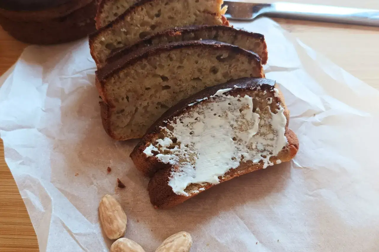 Pokrojony chlebek bananowy z airfryera, posmarowany masłem, obok migdały.