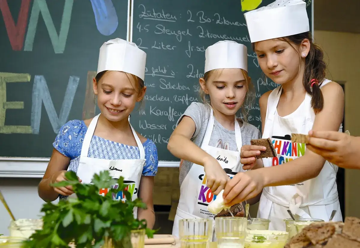 Drei M&auml;dchen mit Kochm&uuml;tzen und Sch&uuml;rzen bereiten in einer K&uuml;che etwas zu. Sie sind konzentriert und haben Spa&szlig;, denn kinder kochen selbst.