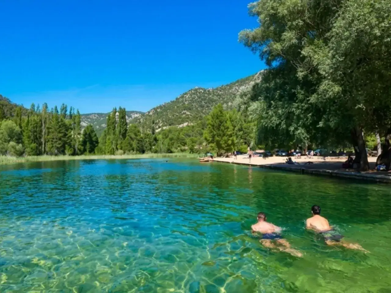 Dos hombres nadan en la cristalina piscina natural Ca&ntilde;amares, rodeada de exuberante vegetaci&oacute;n y monta&ntilde;as.