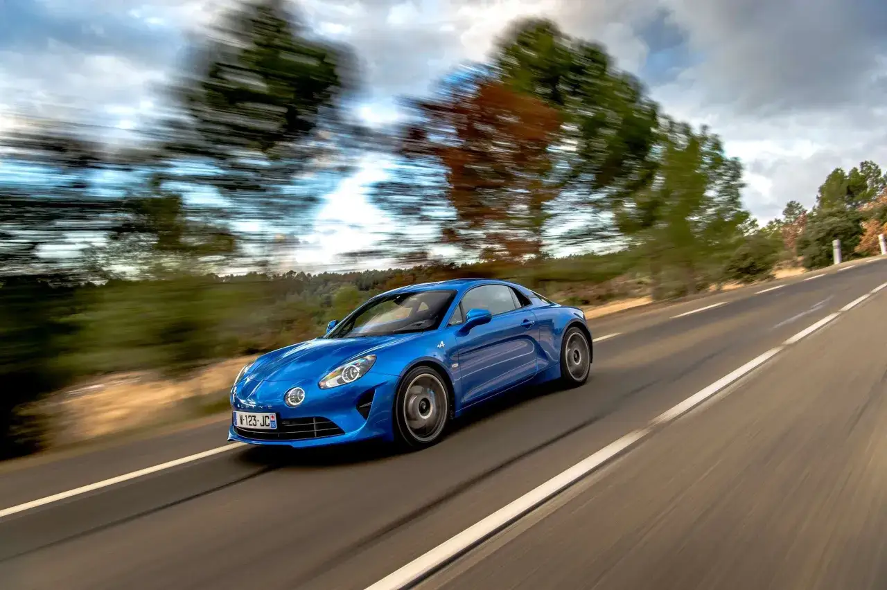 Niebieski Alpine A110 mknie po krętej drodze wśród drzew.