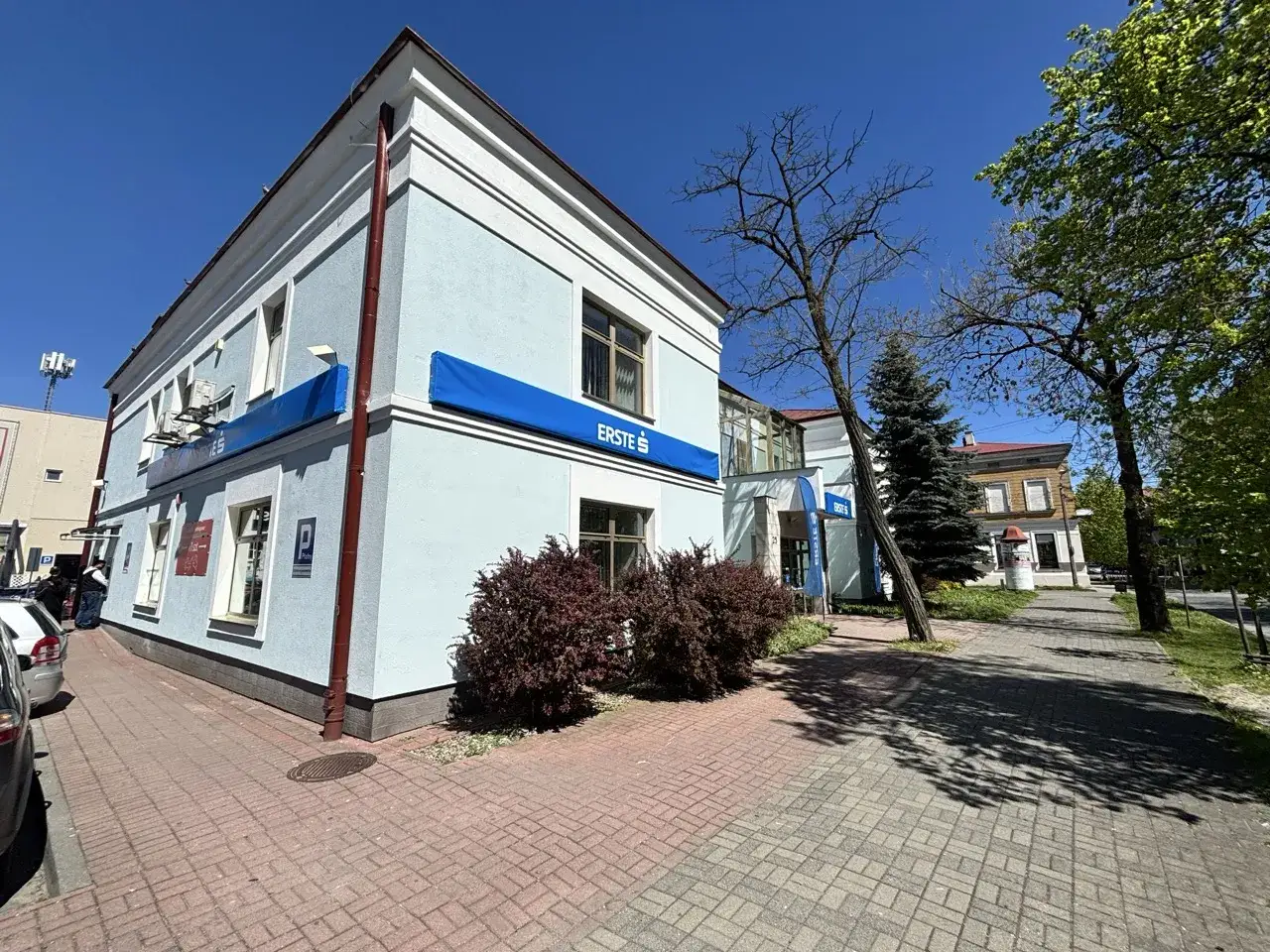 Jasnoniebieski budynek z niebieskim banerem Erste Group. Przed budynkiem chodnik z czerwonej kostki brukowej i krzewy.