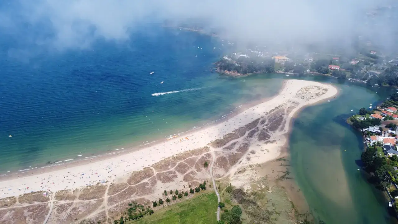 Vista aérea de la playa de Miño con arena dorada, aguas turquesas y casas costeras. Nubes bajas cubren parte del paisaje.