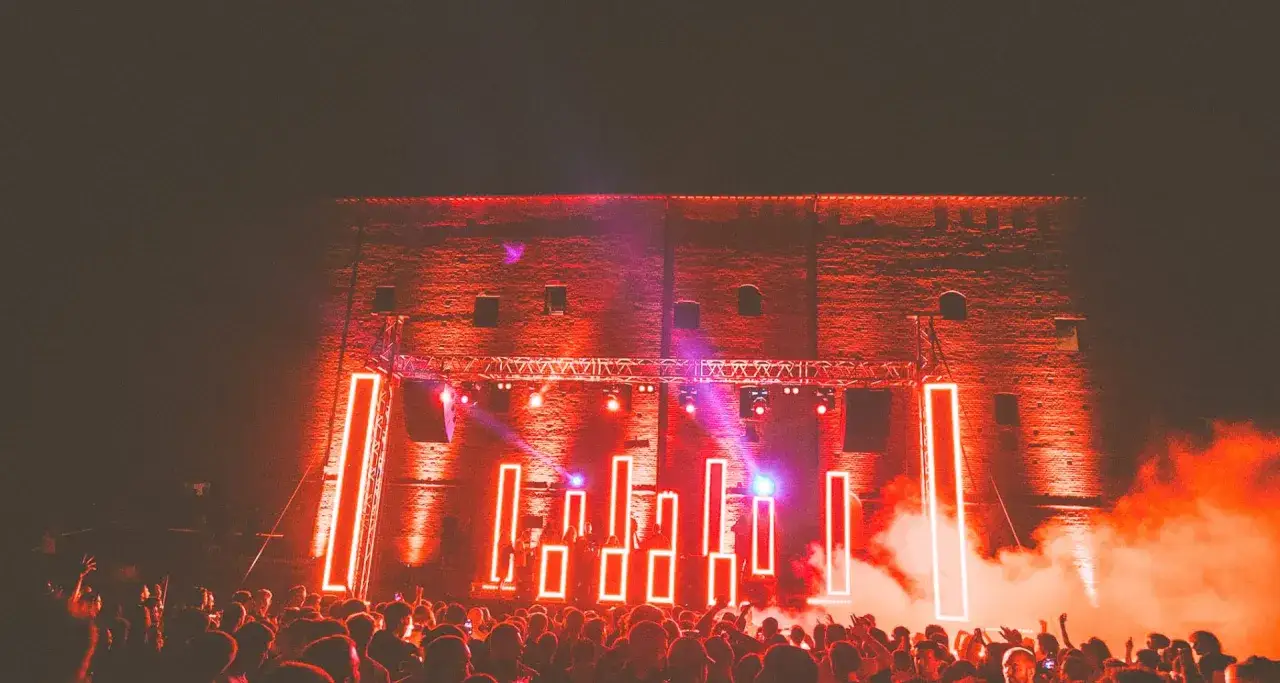 Folla in delirio al **mood festival**, illuminata da luci al neon rosse e fumo denso. La scena è un'esplosione di energia.