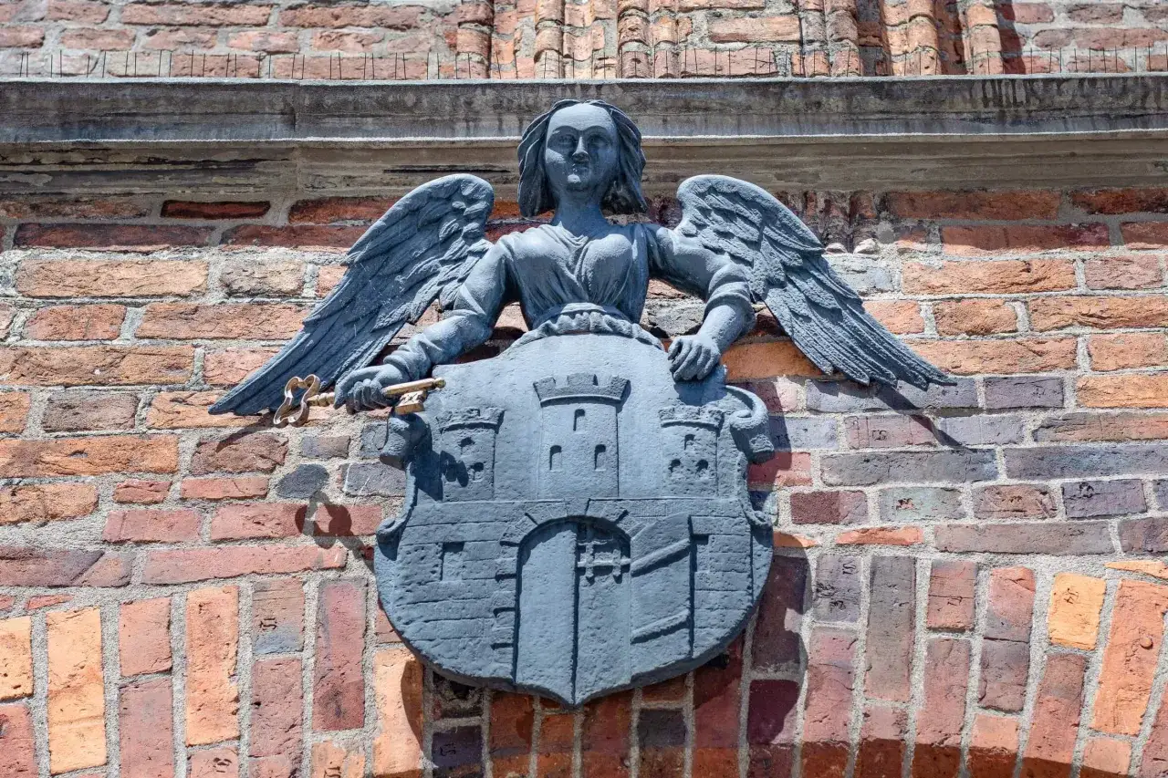 Kamienna rzeźba anioła z kluczem i tarczą z zamkiem, symbolizująca herb Torunia, zdobi ceglaną ścianę.