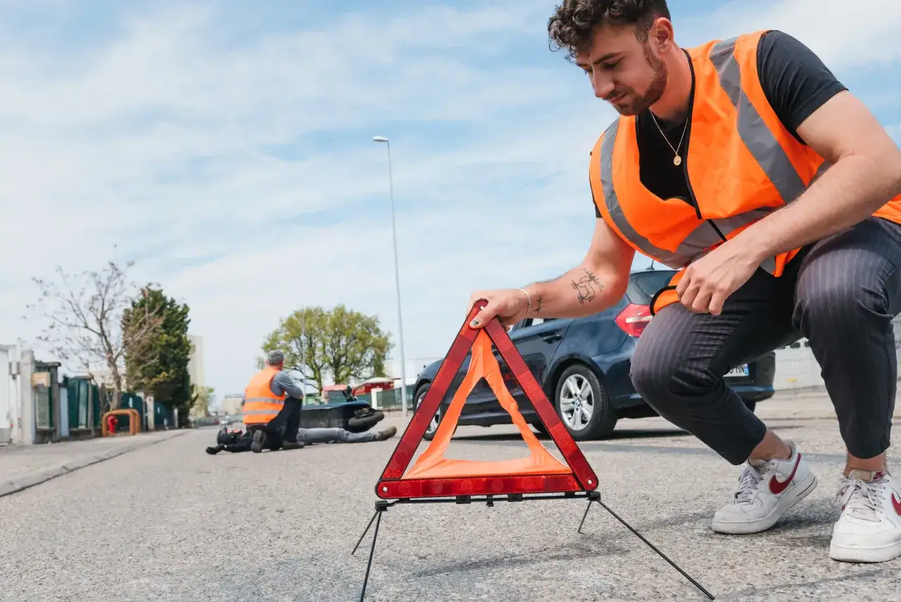 Un homme met en place un triangle de signalisation, un accessoire de sécurité essentiel, près d'une voiture en panne. Un autre homme, portant un gilet de haute visibilité, aide une personne allongée sur la route. Les feux de détresse obligatoires sont ...